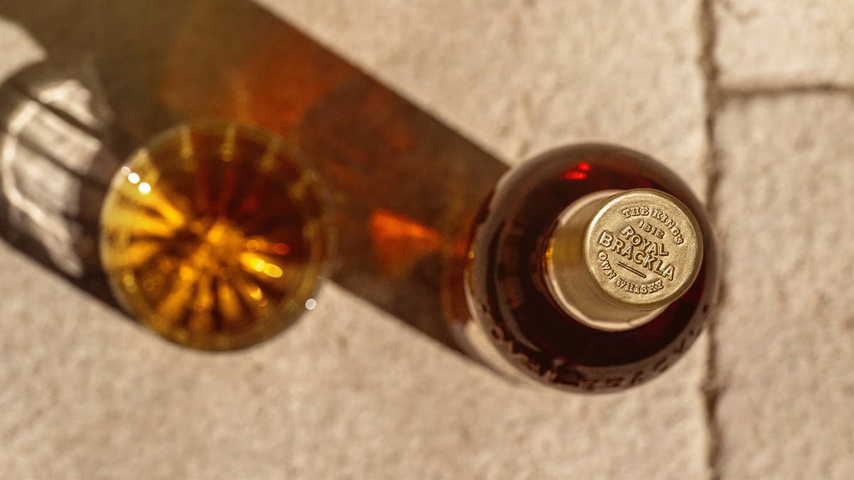 Bottle of Royal Brackla whiskey and tumbler glass lying horizontally on textured stone surface.