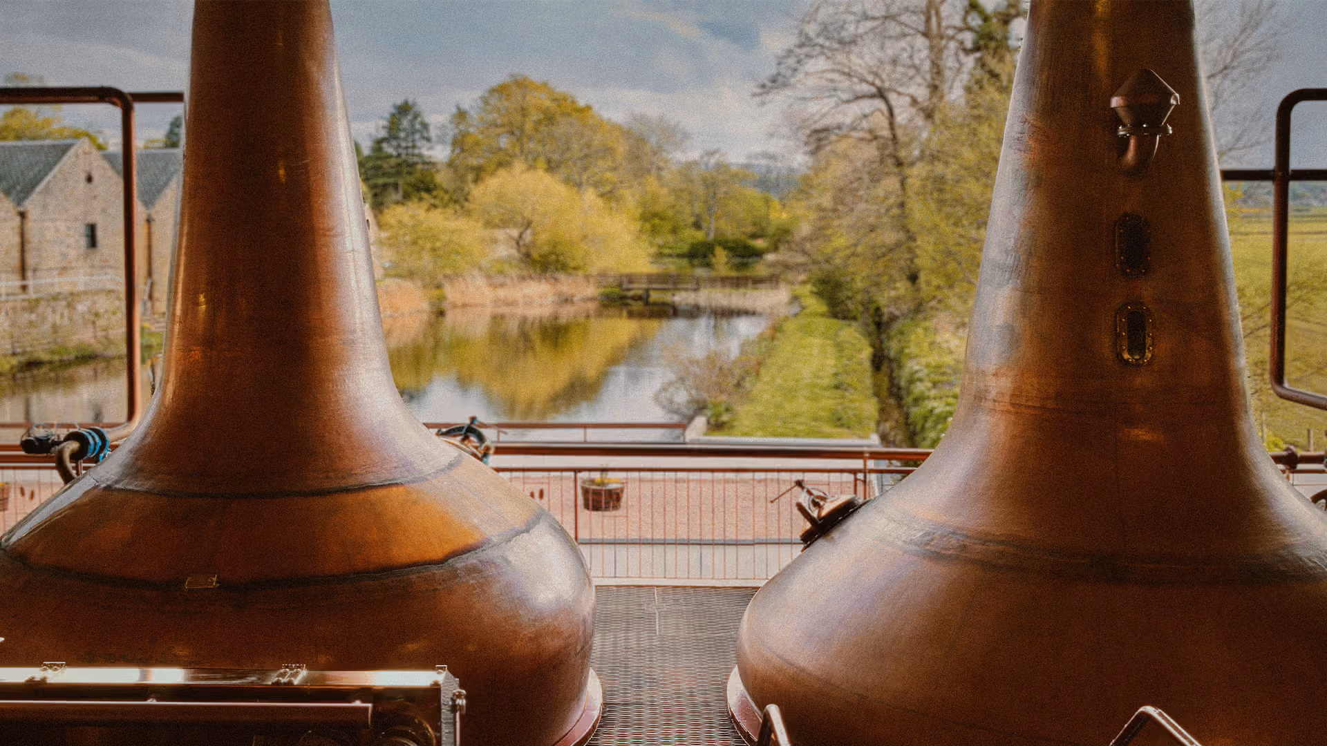 Two large copper pot stills with rounded bases and conical necks framing a river scene with trees, bridge and stone building.